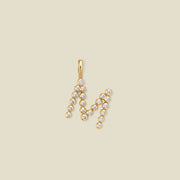 A gold initial pendant letter 'M' set with small clear round gemstones, shown on a solid light beige background.