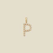 A gold letter 'P' pendant charm embellished with small clear round gemstones, centered on a solid light beige background.