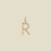 A gold letter 'R' pendant charm embellished with small clear round gemstones, centered on a solid light beige background.