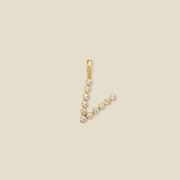A gold initial pendant charm shaped like the letter 'V', adorned with small clear round gemstones, shown on a solid light beige background.