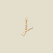 A gold initial pendant 'Y' charm, adorned with small clear round-cut gemstones, set against a solid light beige background.