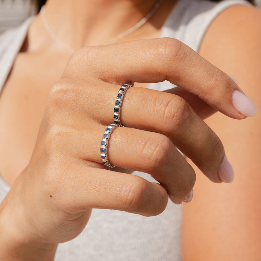 A close-up of a person's hand wearing two silver eternity rings, each with square cut gemstones in either light blue or dark blue.