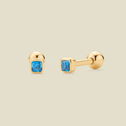 A pair of gold stud earrings with square-cut blue gemstones, shown on a light beige background. One earring is facing forward, and the other is turned to the side to show the post and backing.