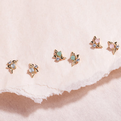 Three pairs of gold stud earrings with prong-set clear stones, prong-set green stones and prong-set pink stones against a neutral paper background.