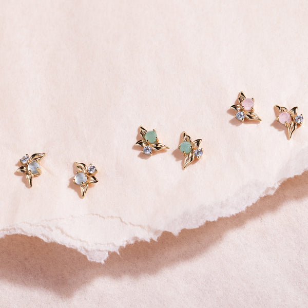 Three pairs of gold stud earrings with prong-set clear stones, prong-set green stones and prong-set pink stones against a neutral paper background.