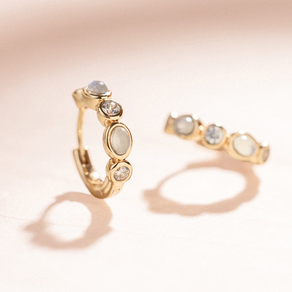 Gold hoop earrings with clear and white bezel-set stones against a neutral background.