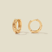 A pair of gold huggie hoop earrings on a light beige background. One earring is shown from the side, displaying a polished outer surface, while the other is shown from the front, showcasing the word 'GEMINI' in raised letters.