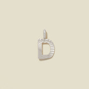 A small silver D-shaped charm with a textured, sunburst striped pattern, shown on a solid light beige background.