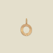 A small gold O-shaped charm with a textured, sunburst striped pattern, shown on a solid light beige background.