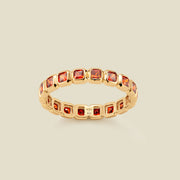 A gold eternity ring featuring a continuous row of square cut red-orange gemstones, shown on a solid light beige background.