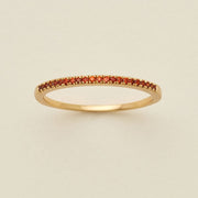 A gold ring featuring a single row of small round-cut red gemstones, photographed on a clean, solid, warm off-white background.