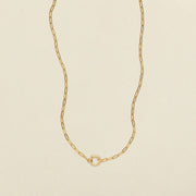 A delicate gold paperclip chain necklace with a small, polished gold ring in the center, displayed on a clean, light beige textured background.
