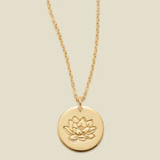 A gold chain necklace with a round gold disc pendant featuring an engraved lotus flower design, displayed against a solid light beige background.