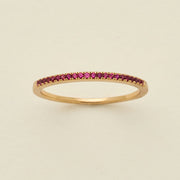 A gold ring featuring a single row of small round-cut dark red gemstones, photographed on a clean, solid, warm off-white background.