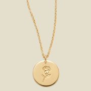 A gold chain necklace with a round gold disc pendant featuring an engraved rose flower design, displayed against a solid light beige background.