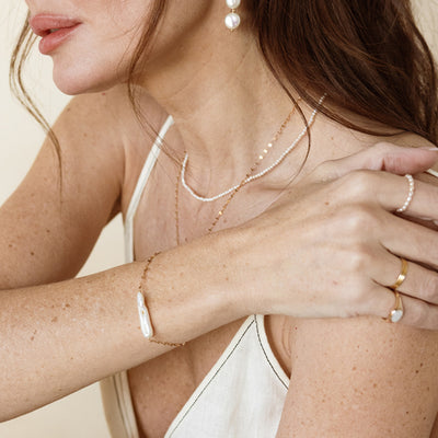 Close-up of a person wearing layered lace chain and pearl strand necklace, a pearl drop earring, a delicate bracelet with a baroque pearl, and gold rings.