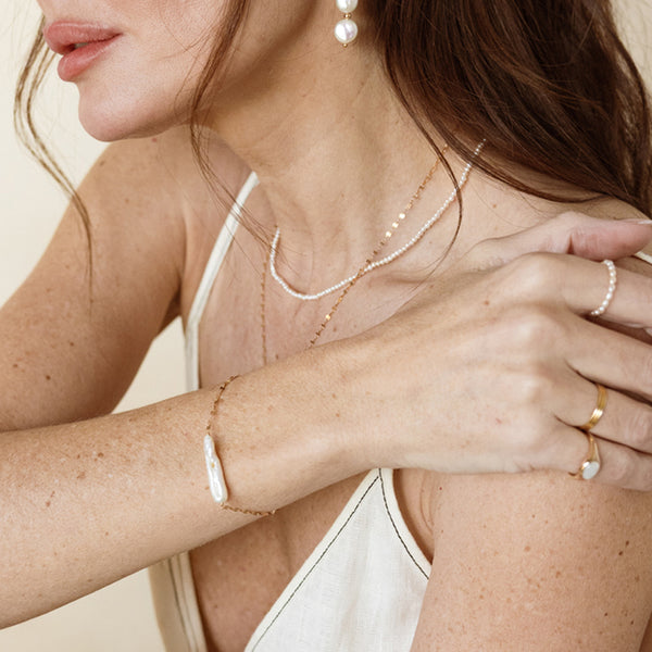 Close-up of a person wearing layered lace chain and pearl strand necklace, a pearl drop earring, a delicate bracelet with a baroque pearl, and gold rings.