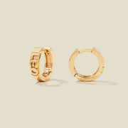 A pair of gold huggie hoop earrings on a light beige background. One earring is shown from the side, displaying a polished outer surface, while the other is shown from the front, showcasing the word 'LEO' in raised letters.