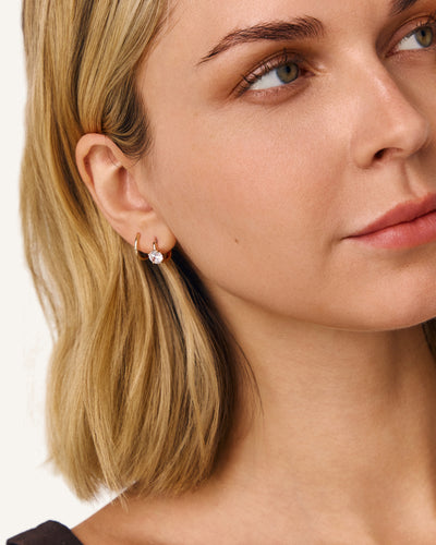 A close-up of a woman with blonde hair wearing a delicate gold hoop earring with a small clear gemstone, shown against a clean white background.