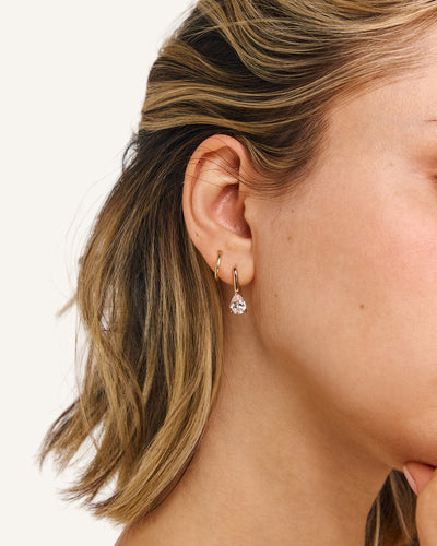A close-up side profile of a woman's ear wearing a small gold huggie hoop earring with a dangling clear teardrop gemstone, against a plain light background.