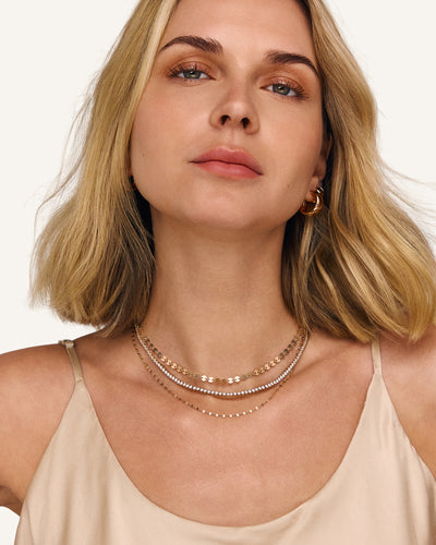 A close-up of a blonde woman wearing three layered necklaces with gold chains, paired with a beige camisole top against a clean white background.
