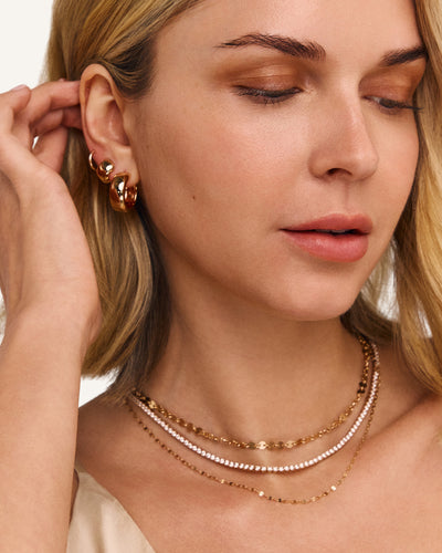 Close-up of a woman wearing layered gold necklaces and gold hoop earrings, with her hand near her ear against a clean white background.
