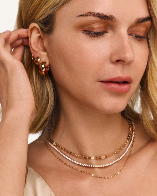 Close-up of a woman wearing layered gold necklaces and gold hoop earrings, with her hand near her ear against a clean white background.