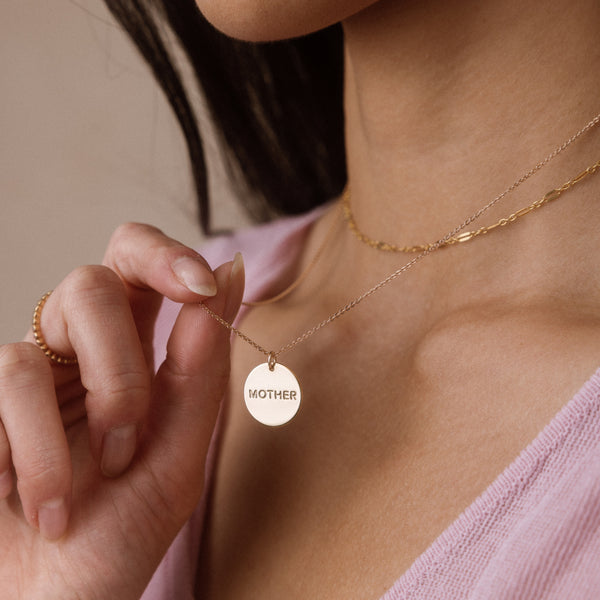 A close-up lifestyle shot of a woman wearing a delicate gold chain necklace with a round disc pendant engraved with the word 'MOTHER'. She is wearing a light pink top, and the background is a soft, neutral beige.
