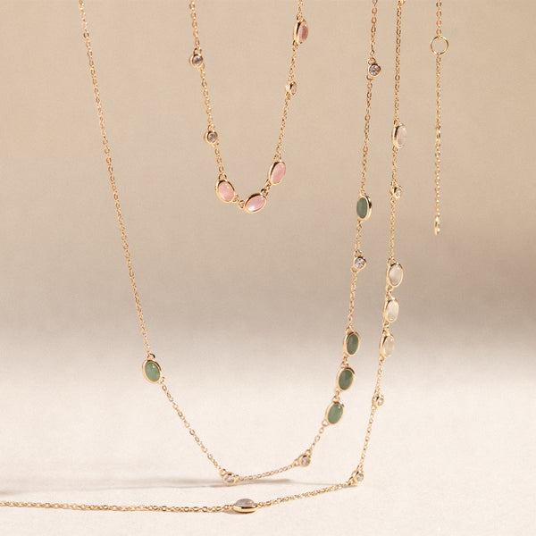 Three gold necklaces suspended against a plain beige background. Pink bezel-set stone necklace, green bezel-set necklace and a white bezel-set stone necklace.