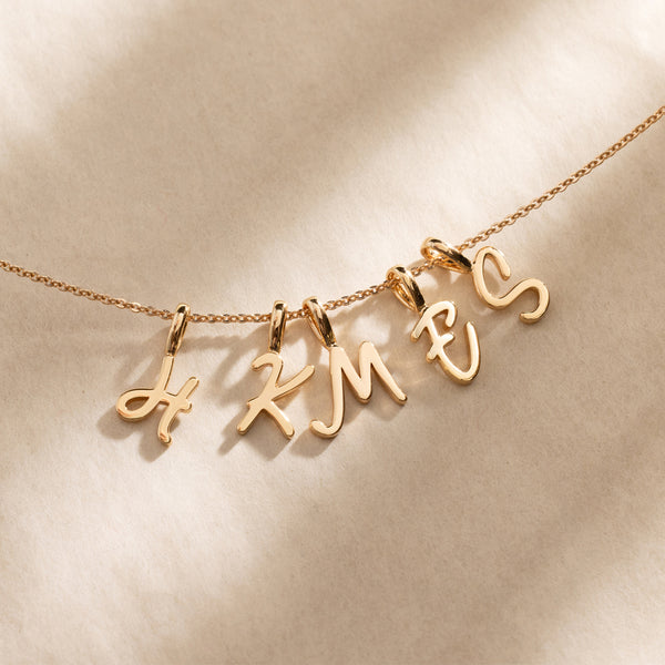 Close-up of a gold chain necklace featuring multiple individual cursive initial charms spaced along the chain.