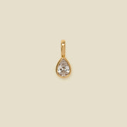 A gold teardrop-shaped pendant with a clear faceted gemstone, centered on a solid beige background.
