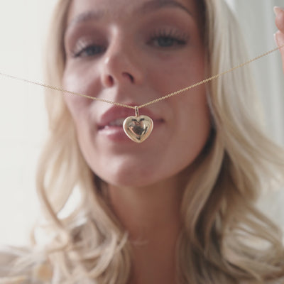 A short video of a blonde woman holding and wearing a delicate gold chain necklace with a large polished heart charm.