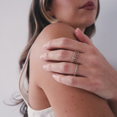A short video of a model in a beige tank top wearing multiple stacked gold rings, each with a different color of square cut gemstones in a single continuous row.