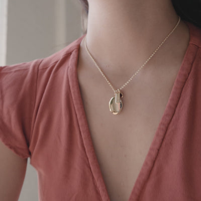 A short video of a close-up of a person in a red t-shirt wearing a gold chain necklace with an oval locket charm, a small rectangular bar engraved with the word 'LOVE', and a square red gemstone charm.