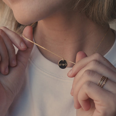 A short video of a blonde woman in a white t-shirt wearing a delicate gold pendant chain necklace with a round disc engraved with the letters 'K' and 'J' separated by a heart.