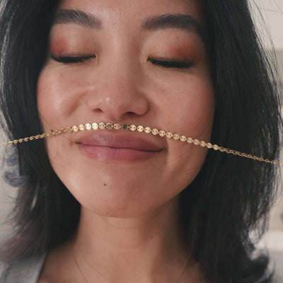 Short video of a model wearing and playing with a gold chain necklace with flat round linked discs, paired with a grey long sleeve knit shirt and set in a neutral colored bedroom.