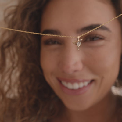 A short video of a model holding a delicate pendant chain with a gold hummingbird charm in front of their face, then wearing the necklace layered with a dainty pearl necklace.