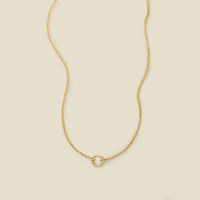 A gold circular link gold chain necklace with a round ring in the center, shown on a solid light beige background.