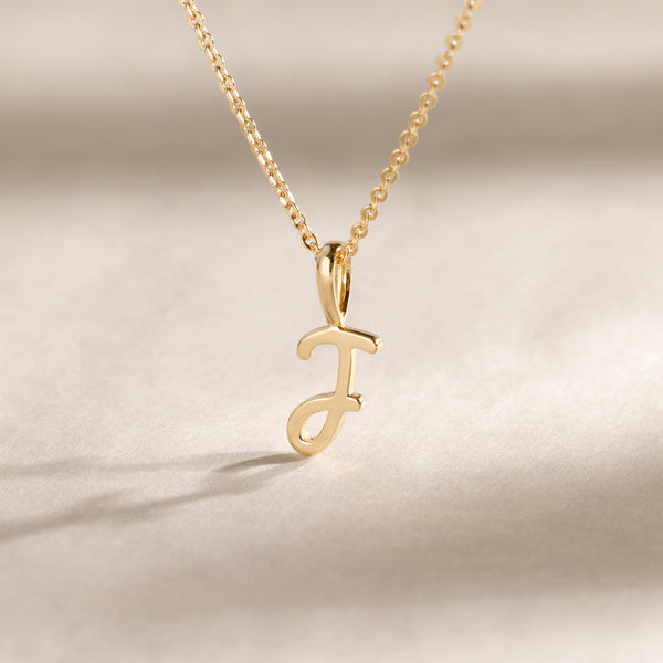 Close-up of a gold chain necklace featuring a polished cursive initial “J” pendant resting on a neutral surface.