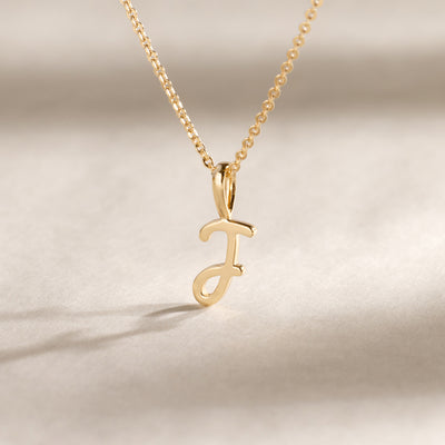 Close-up of a gold chain necklace featuring a polished cursive initial “J” pendant resting on a neutral surface.