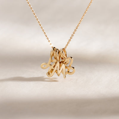 Close-up of a gold chain necklace with multiple cursive initial charms clustered together, against a neutral background.