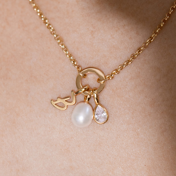 A close-up of a gold necklace worn on a person’s neck. It features a link lock holding three small charms: a cursive initial charm, a smooth oval pearl, and a teardrop-shaped clear gemstone.