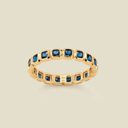A gold eternity ring featuring a continuous row of square cut dark blue gemstones, shown on a solid light beige background.